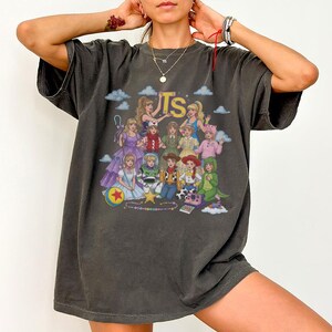 May include: A charcoal gray short-sleeve t-shirt featuring a graphic print of various animated characters and the letters TS in yellow. The design includes multiple figures in colorful outfits, a ball with a red star, and small blue clouds.