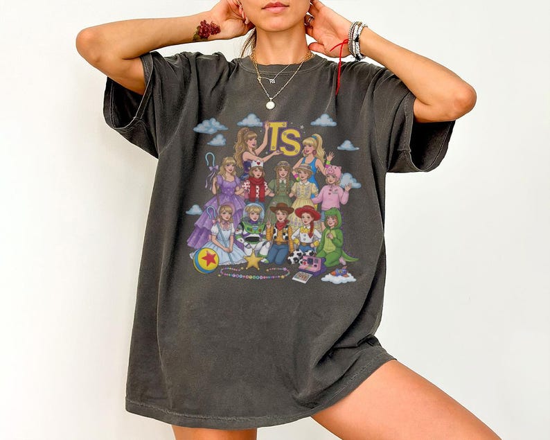 May include: A charcoal gray short-sleeve t-shirt featuring a graphic print of various animated characters and the letters TS in yellow. The design includes multiple figures in colorful outfits, a ball with a red star, and small blue clouds.
