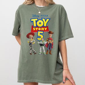 Op de afbeelding: Een olijfgroen T-shirt met korte mouwen en een grafische print van het Disney Pixar Toy Story 5-logo, met de personages Jessie, Buzz Lightyear en Woody onder de tekst. Het shirt heeft een ronde hals en een ruimvallend ontwerp.