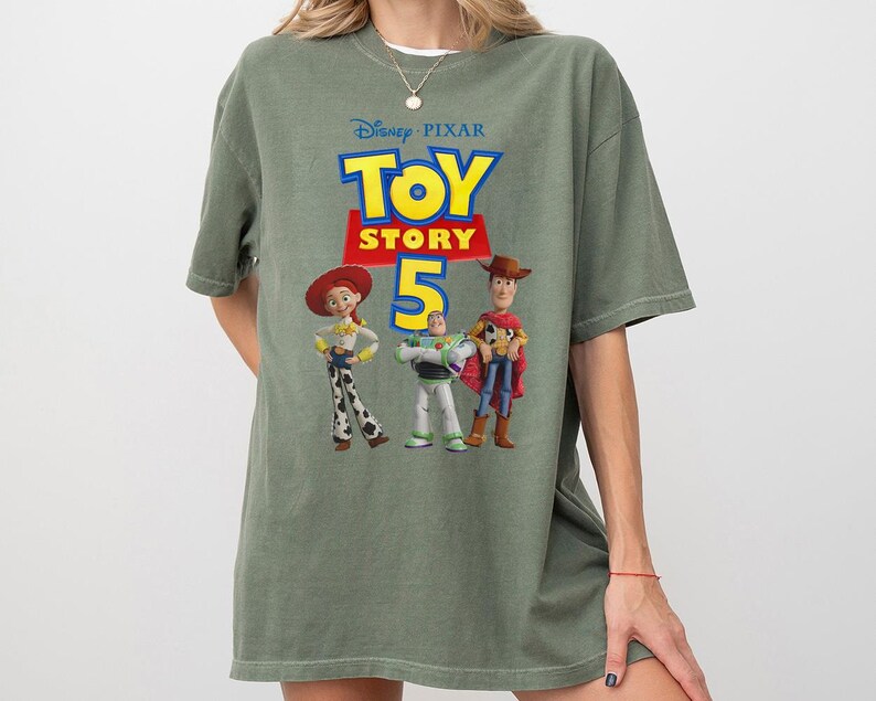 Op de afbeelding: Een olijfgroen T-shirt met korte mouwen en een grafische print van het Disney Pixar Toy Story 5-logo, met de personages Jessie, Buzz Lightyear en Woody onder de tekst. Het shirt heeft een ronde hals en een ruimvallend ontwerp.