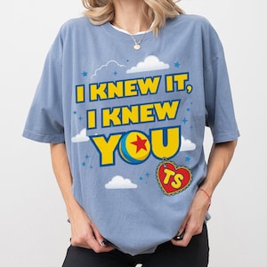 Pode incluir: Uma t-shirt de manga curta em azul poeirento com uma impress&atilde;o gr&aacute;fica com o texto amarelo I KNEW IT, I KNEW YOU. O design inclui uma bola azul, amarela, azul e vermelha, nuvens brancas, pequenas estrelas azuis e uma etiqueta vermelha em forma de cora&ccedil;&atilde;o com as letras TS.