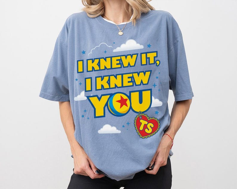 Pode incluir: Uma t-shirt de manga curta em azul poeirento com uma impress&atilde;o gr&aacute;fica com o texto amarelo I KNEW IT, I KNEW YOU. O design inclui uma bola azul, amarela, azul e vermelha, nuvens brancas, pequenas estrelas azuis e uma etiqueta vermelha em forma de cora&ccedil;&atilde;o com as letras TS.