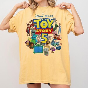 May include: A pale yellow short-sleeve t-shirt featuring a colorful graphic print of various Toy Story characters and the text Disney Pixar Toy Story 5. The design includes figures like Woody, Buzz Lightyear, Jessie, and others arranged around the large blue number five.