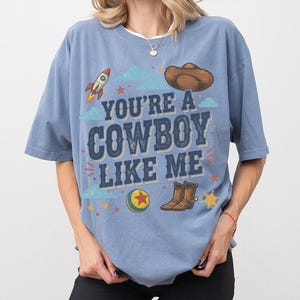 May include: A blue short-sleeve t-shirt featuring the text "YOU'RE A COWBOY LIKE ME" in dark blue letters. The graphic includes a brown cowboy hat, a rocket, a colourful ball with a red star, and brown cowboy boots, surrounded by small stars.