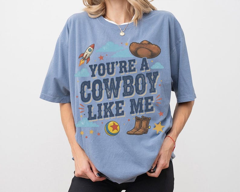 May include: A blue short-sleeve t-shirt featuring the text "YOU'RE A COWBOY LIKE ME" in dark blue letters. The graphic includes a brown cowboy hat, a rocket, a colourful ball with a red star, and brown cowboy boots, surrounded by small stars.