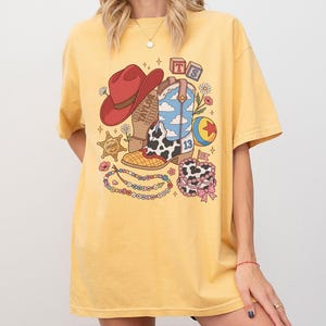 May include: A yellow short-sleeve t-shirt featuring a graphic print of a red cowboy hat, a cow-print boot with a blue sky pattern, a sheriff star, a colourful ball, a beaded necklace, and a cake. The text TS and TAYLOR are visible in the design.