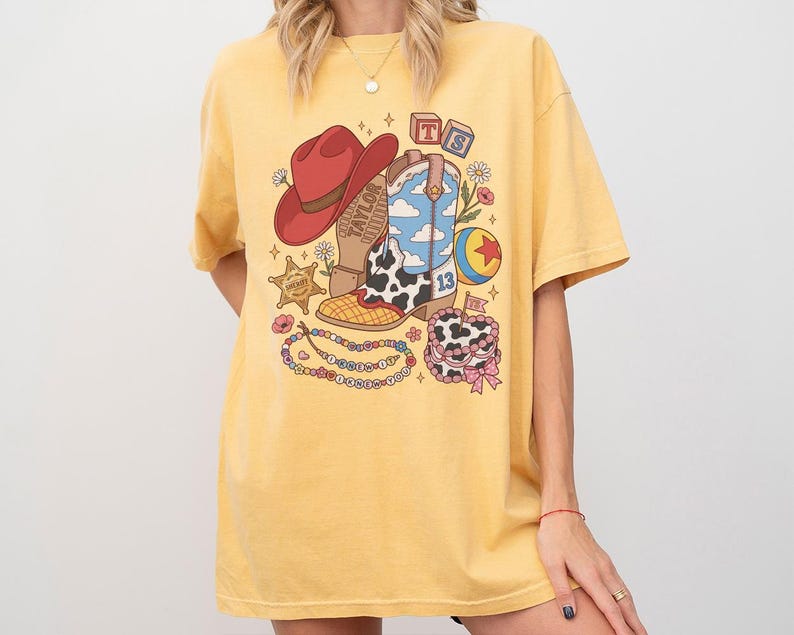 May include: A yellow short-sleeve t-shirt featuring a graphic print of a red cowboy hat, a cow-print boot with a blue sky pattern, a sheriff star, a colourful ball, a beaded necklace, and a cake. The text TS and TAYLOR are visible in the design.