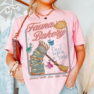 May include: Pink t-shirt featuring a whimsical design for "Fauna Bakery" with a cake, fairies, and the text "Once Upon a Time." The design includes the text "Baked With Love And Mag" and "Since 1959."