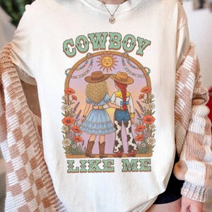 Puede incluir: Una camiseta color crema con un estampado gr&aacute;fico de dos figuras con sombreros de vaquero en un campo de flores bajo un sol. El texto "COWBOY" aparece sobre la ilustraci&oacute;n y "LIKE ME" debajo en una tipograf&iacute;a verde de estilo retro.