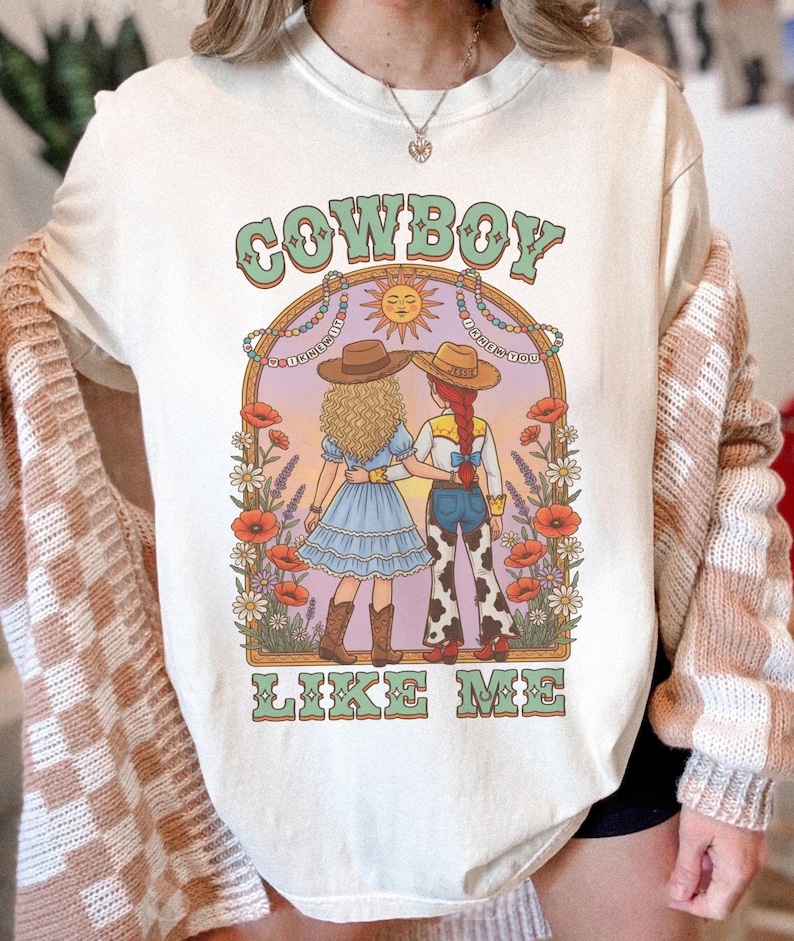 Puede incluir: Una camiseta color crema con un estampado gr&aacute;fico de dos figuras con sombreros de vaquero en un campo de flores bajo un sol. El texto "COWBOY" aparece sobre la ilustraci&oacute;n y "LIKE ME" debajo en una tipograf&iacute;a verde de estilo retro.