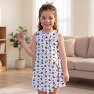 May include: A sleeveless white dress featuring an all-over print of green three-eyed aliens, red and blue balloons, and small planets. The dress has a round neckline and a side tie detail at the waist, creating a wrap-style skirt effect.