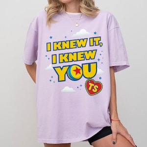 Pode incluir: Uma t-shirt roxo-claro com o texto I KNEW IT, I KNEW YOU em letras amarelas em negrito com contornos azuis. O design inclui uma bola colorida com uma estrela vermelha, pequenas nuvens brancas, estrelas azuis e uma etiqueta vermelha em forma de cora&ccedil;&atilde;o com as letras TS.