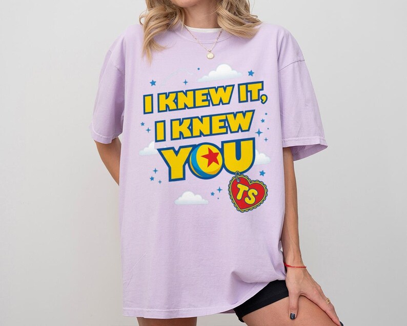 Pode incluir: Uma t-shirt roxo-claro com o texto I KNEW IT, I KNEW YOU em letras amarelas em negrito com contornos azuis. O design inclui uma bola colorida com uma estrela vermelha, pequenas nuvens brancas, estrelas azuis e uma etiqueta vermelha em forma de cora&ccedil;&atilde;o com as letras TS.