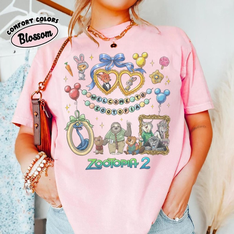 May include: Pink t-shirt featuring a colorful graphic design with characters and text from the movie "Zootopia 2". The design includes framed images, balloons, and the words "WELCOME TO ZOOTOPIA". The shirt also has the words "COMFORT COLORS" and "Blossom".