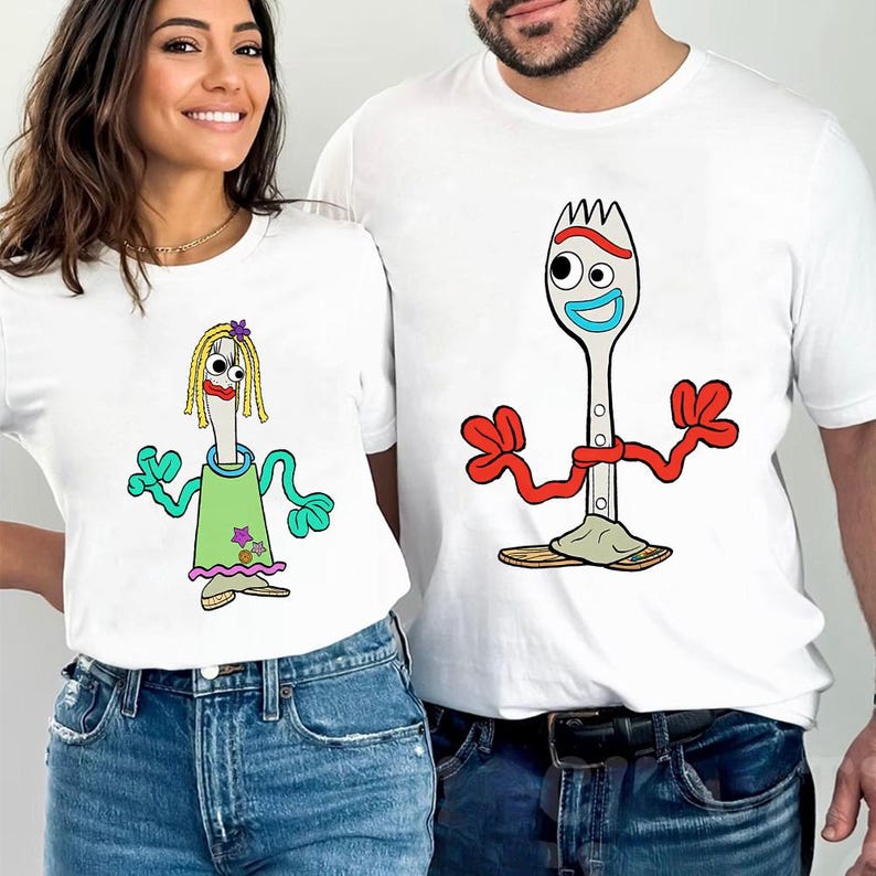 May include: Two white t-shirts featuring cartoon characters. The left shirt displays a spork character with yellow yarn hair, red lips, and a green dress. The right shirt displays a spork character with red pipe cleaner arms and blue mouth.