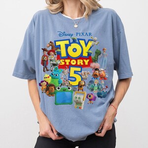 May include: A dusty blue short-sleeve t-shirt featuring a large graphic print of the Disney Pixar Toy Story 5 logo in yellow and red, surrounded by a colorful collage of various animated characters from the film franchise including toys.