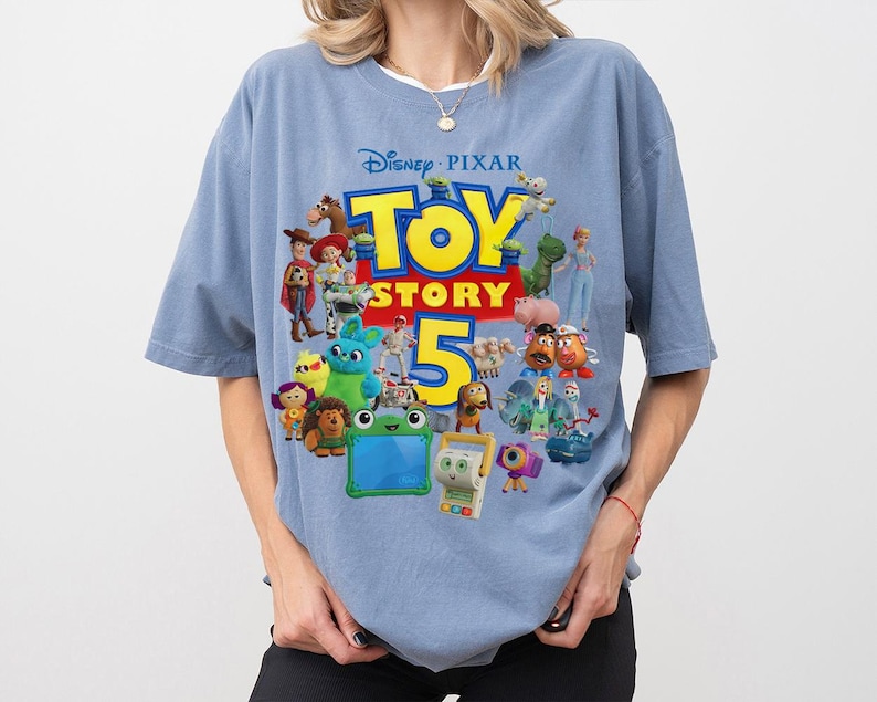 May include: A dusty blue short-sleeve t-shirt featuring a large graphic print of the Disney Pixar Toy Story 5 logo in yellow and red, surrounded by a colorful collage of various animated characters from the film franchise including toys.