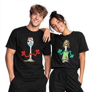 May include: Two black t-shirts featuring cartoon character graphics. The left shirt displays a white spork character with red pipe cleaner arms, googly eyes, and a blue mouth. The right shirt shows a spork character with yellow yarn hair, a green dress, and red lips.