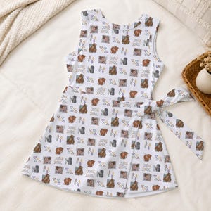 May include: A sleeveless white dress featuring a repeating pattern of brown bears, grey wolves, and small geometric triangles. The garment includes a wrap-style skirt section with a fabric tie closure on the right side, creating a layered look.