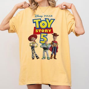 Op de afbeelding: Een lichtgeel T-shirt met korte mouwen en een grafische print van het Disney Pixar Toy Story 5-logo, met de personages Jessie, Buzz Lightyear en Woody onder de tekst. Het T-shirt heeft een ronde hals en een losse, relaxte pasvorm.