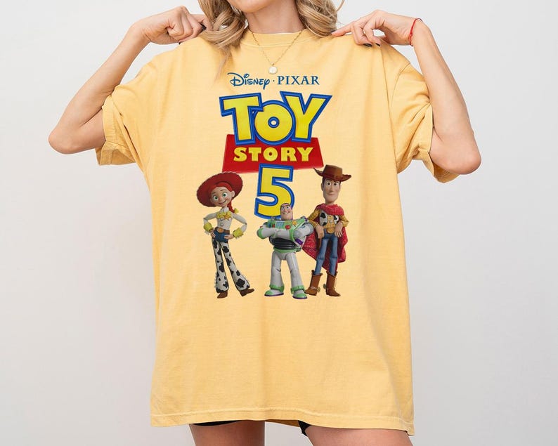 Op de afbeelding: Een lichtgeel T-shirt met korte mouwen en een grafische print van het Disney Pixar Toy Story 5-logo, met de personages Jessie, Buzz Lightyear en Woody onder de tekst. Het T-shirt heeft een ronde hals en een losse, relaxte pasvorm.