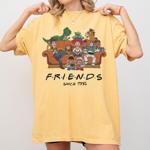 K&ouml;nnte beinhalten: Ein gelbes T-Shirt mit einem Grafikdruck verschiedener animierter Figuren, die auf einem braunen Sofa sitzen. Unter dem Sofa steht der Text F.R.I.E.N.D.S SINCE 1995 in schwarzer Schrift mit bunten Punkten zwischen den Buchstaben des Wortes FRIENDS.