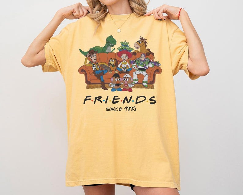 K&ouml;nnte beinhalten: Ein gelbes T-Shirt mit einem Grafikdruck verschiedener animierter Figuren, die auf einem braunen Sofa sitzen. Unter dem Sofa steht der Text F.R.I.E.N.D.S SINCE 1995 in schwarzer Schrift mit bunten Punkten zwischen den Buchstaben des Wortes FRIENDS.