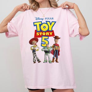 Op de afbeelding: Een lichtroze T-shirt met korte mouwen en een grafische print van het Disney Pixar Toy Story 5-logo, met de personages Jessie, Buzz Lightyear en Woody onder de tekst. Het shirt heeft een ronde hals en een ruime, losse pasvorm.