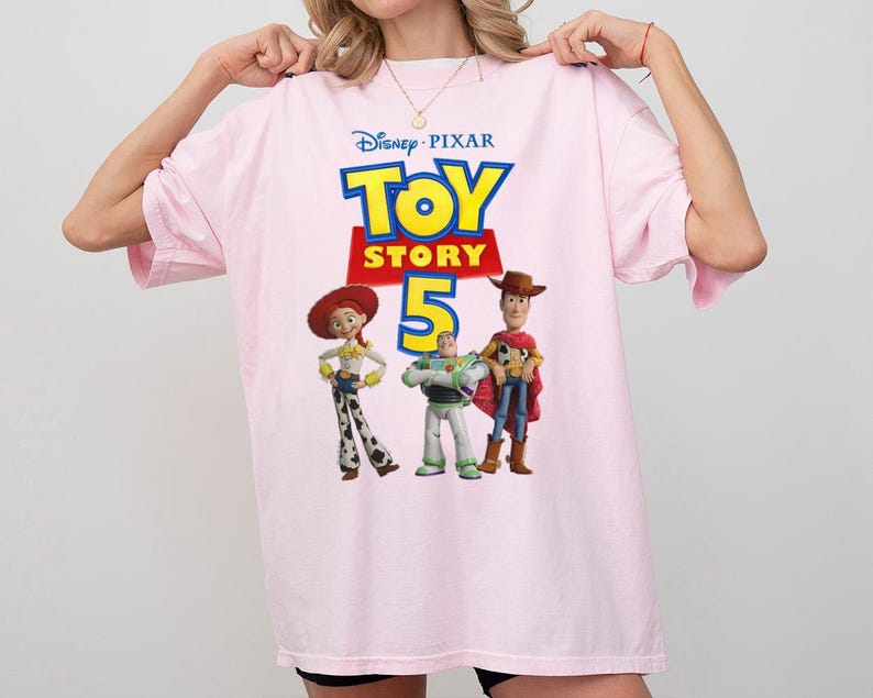 Op de afbeelding: Een lichtroze T-shirt met korte mouwen en een grafische print van het Disney Pixar Toy Story 5-logo, met de personages Jessie, Buzz Lightyear en Woody onder de tekst. Het shirt heeft een ronde hals en een ruime, losse pasvorm.