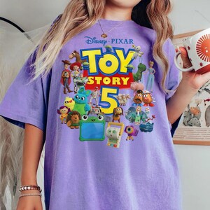 May include: A light purple t-shirt featuring a large graphic print of the Disney Pixar Toy Story 5 logo surrounded by various animated characters including Woody, Buzz Lightyear, Jessie, Rex, Hamm, Mr. Potato Head, and other colorful toy figures.