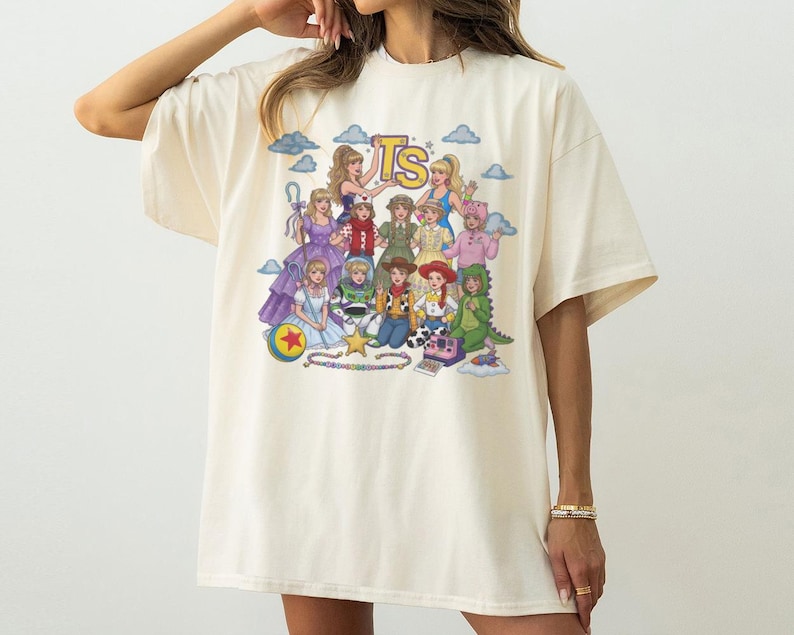 May include: An off-white short-sleeve t-shirt featuring a colorful graphic print of various animated characters dressed in costumes. The design includes the letters TS at the top, a yellow ball with a red star, a star shape, and a camera icon.