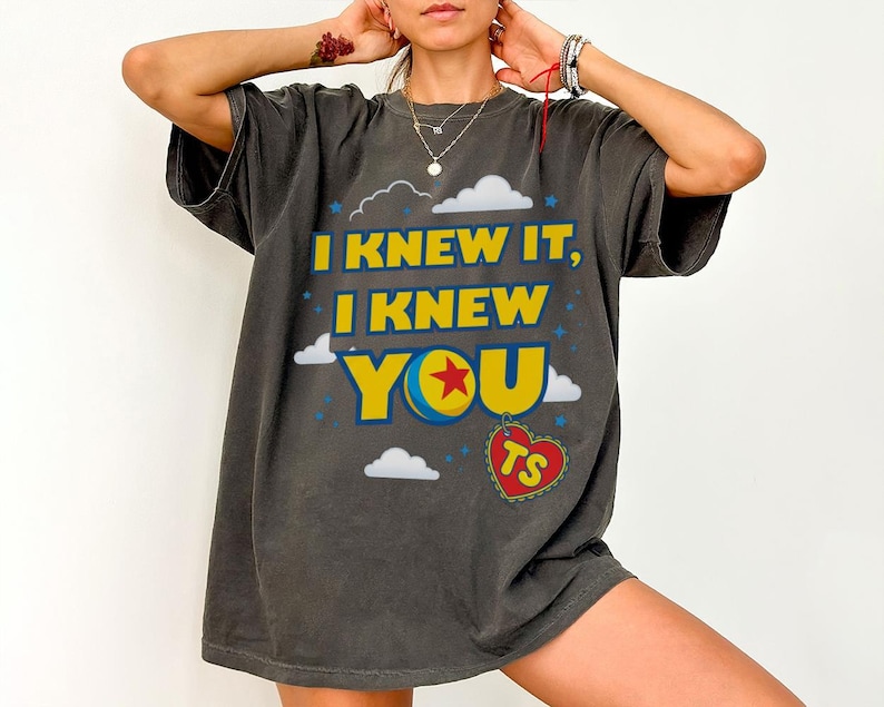 Pode incluir: Uma t-shirt de manga curta cinza-carv&atilde;o com o texto amarelo I KNEW IT, I KNEW YOU, um gr&aacute;fico de bola colorida substituindo a letra O e uma etiqueta vermelha em forma de cora&ccedil;&atilde;o com as letras TS pendurada abaixo, rodeada por nuvens brancas.