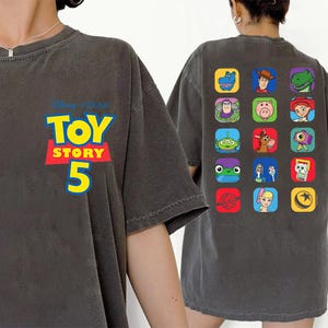 May include: A charcoal gray t-shirt featuring the Disney Pixar Toy Story 5 logo on the front left chest. The back of the shirt displays a grid of fifteen colorful square icons, each depicting a different character from the Toy Story film series.
