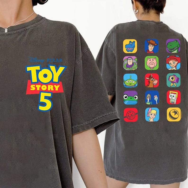May include: A charcoal gray t-shirt featuring the Disney Pixar Toy Story 5 logo on the front left chest. The back of the shirt displays a grid of fifteen colorful square icons, each depicting a different character from the Toy Story film series.