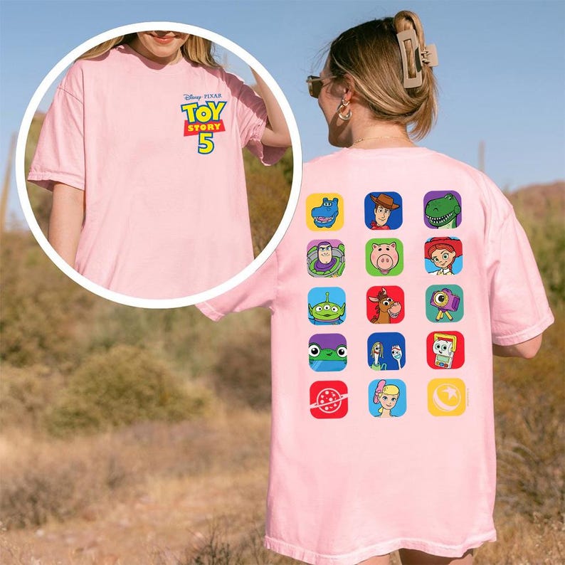 May include: A light pink short-sleeve t-shirt featuring a small Disney Pixar Toy Story 5 logo on the front chest. The back displays a grid of fifteen colorful square icons depicting various animated characters and symbols from the film series.