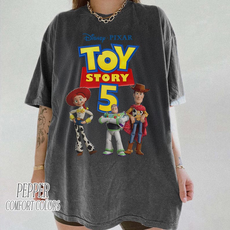 Op de afbeelding: Een antracietgrijs t-shirt met korte mouwen en een grafische print van het Disney Pixar Toy Story 5-logo met de personages Jessie, Buzz Lightyear en Woody. Het shirt heeft een relaxte pasvorm en bevat de tekst Pepper Comfort Colors.