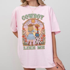 Puede incluir: Una camiseta de color rosa claro con un estampado gr&aacute;fico de dos figuras con sombreros de vaquero dentro de un arco. El texto "COWBOY" aparece sobre el arco y "LIKE ME" debajo. La ilustraci&oacute;n incluye flores, un sol y un paisaje des&eacute;rtico.
