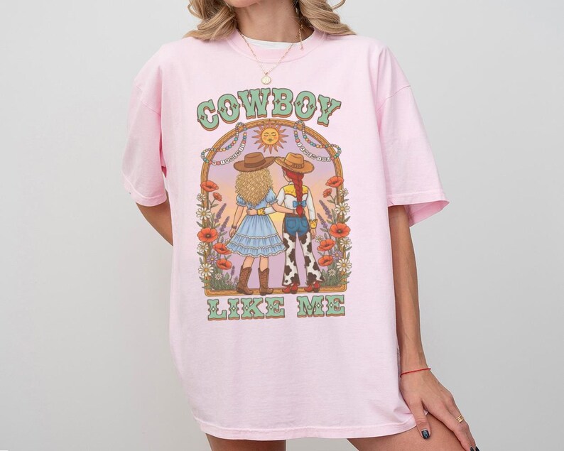 Puede incluir: Una camiseta de color rosa claro con un estampado gr&aacute;fico de dos figuras con sombreros de vaquero dentro de un arco. El texto "COWBOY" aparece sobre el arco y "LIKE ME" debajo. La ilustraci&oacute;n incluye flores, un sol y un paisaje des&eacute;rtico.