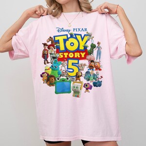 May include: A light pink t-shirt featuring a colorful graphic print of the Disney Pixar Toy Story 5 logo. The design includes various animated characters from the film franchise arranged around the large yellow and red text on the front of the shirt.
