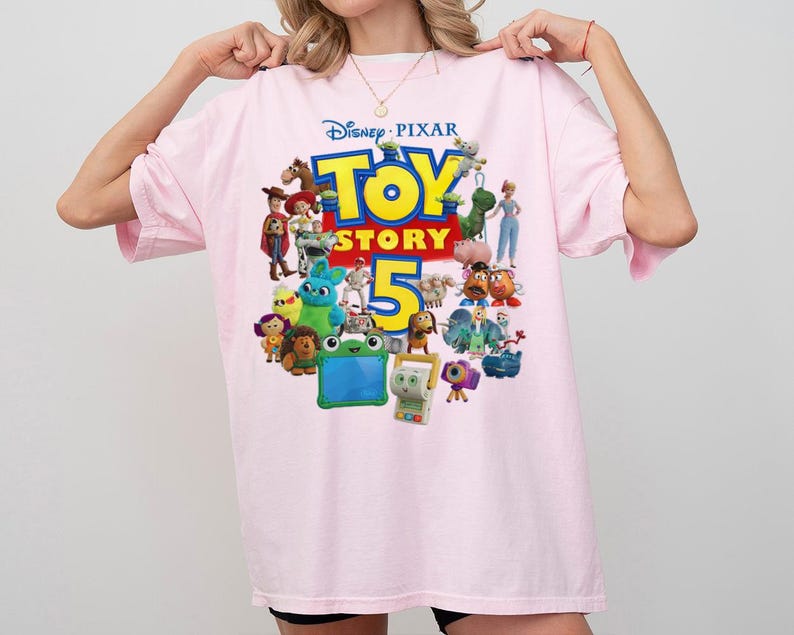 May include: A light pink t-shirt featuring a colorful graphic print of the Disney Pixar Toy Story 5 logo. The design includes various animated characters from the film franchise arranged around the large yellow and red text on the front of the shirt.