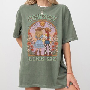 Puede incluir: Una camiseta verde oliva con un estampado gr&aacute;fico de dos figuras con sombreros de vaquero bajo un arco con el texto COWBOY LIKE ME. La ilustraci&oacute;n incluye flores, un sol y las figuras visten vestidos y pantalones con estampado de vaca.