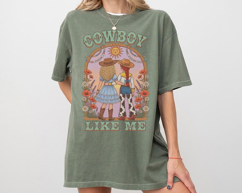 Puede incluir: Una camiseta verde oliva con un estampado gr&aacute;fico de dos figuras con sombreros de vaquero bajo un arco con el texto COWBOY LIKE ME. La ilustraci&oacute;n incluye flores, un sol y las figuras visten vestidos y pantalones con estampado de vaca.