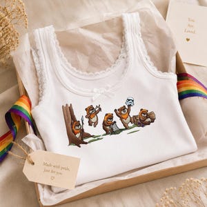 May include: A white ribbed tank top with lace trim along the neckline and straps, featuring a small center bow. The front displays a graphic of five brown Ewok characters in various poses, including one holding a spear and one holding a helmet.