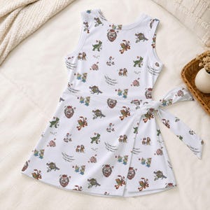 May include: A white sleeveless wrap dress featuring an all-over print of various cartoon characters from Toy Story wearing holiday accessories like Santa hats and scarves, with a fabric tie closure at the right side of the waist.