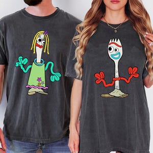 May include: Two charcoal gray t-shirts featuring cartoon characters. The left shirt displays a white utensil character with yellow yarn hair, a green dress, and teal arms. The right shirt shows a white spork character with red arms and a blue mouth.