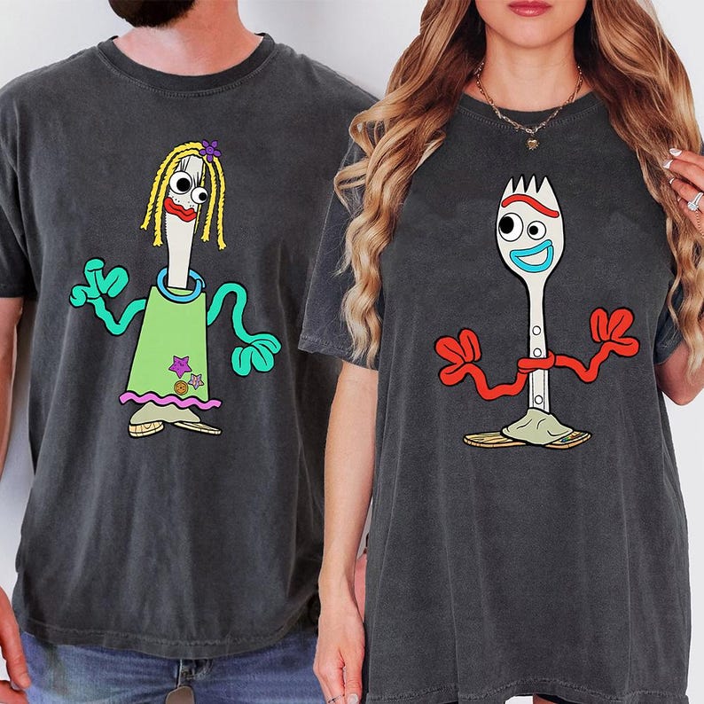 May include: Two charcoal gray t-shirts featuring cartoon characters. The left shirt displays a white utensil character with yellow yarn hair, a green dress, and teal arms. The right shirt shows a white spork character with red arms and a blue mouth.