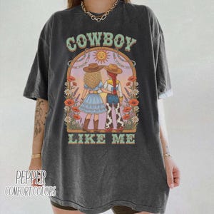 Puede incluir: Una camiseta gris carb&oacute;n de manga corta con un estampado gr&aacute;fico de dos figuras con sombreros de vaquero bajo un arco floral. El texto COWBOY LIKE ME est&aacute; impreso en verde claro. El dise&ntilde;o incluye un sol y diversas flores.