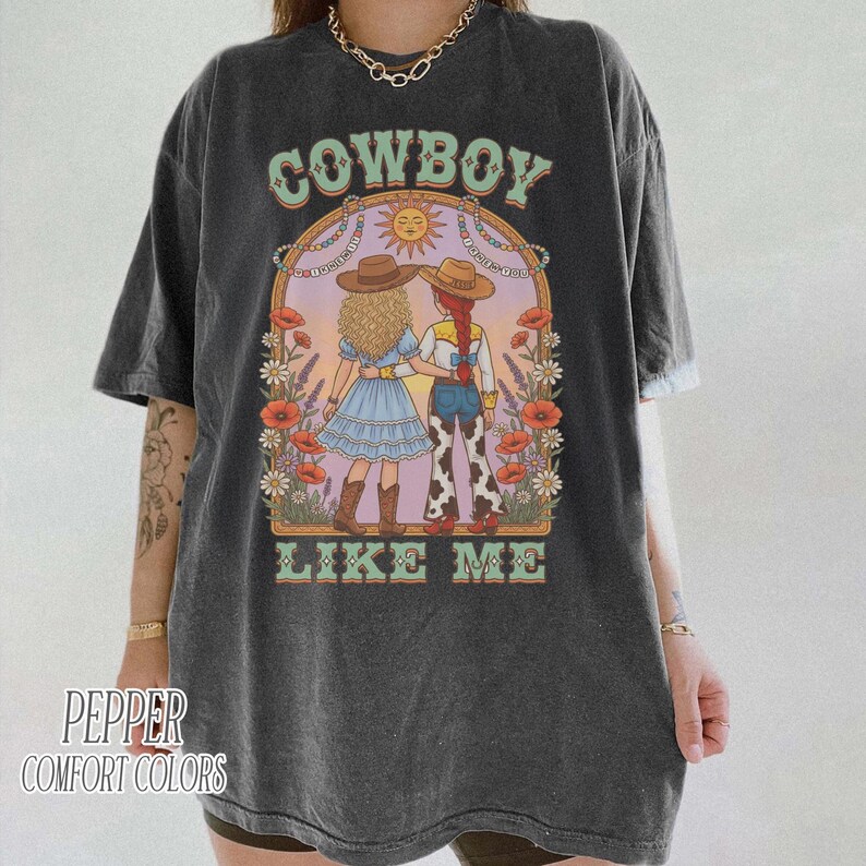 Puede incluir: Una camiseta gris carb&oacute;n de manga corta con un estampado gr&aacute;fico de dos figuras con sombreros de vaquero bajo un arco floral. El texto COWBOY LIKE ME est&aacute; impreso en verde claro. El dise&ntilde;o incluye un sol y diversas flores.