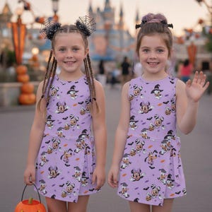 Może przedstawiać: Dwie bezrękawowe sukienki w kolorze lawendowym z halloweenowym nadrukiem przedstawiającym postacie z kresk&oacute;wek w kapeluszach czarownic, dynie i nietoperze. Sukienki mają kopertowy d&oacute;ł z wiązaniem z boku oraz okrągły dekolt.