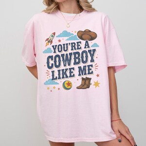 May include: A light pink t-shirt featuring the dark blue text "YOU'RE A COWBOY LIKE ME" printed on the chest. The graphic includes a brown cowboy hat, a rocket ship, a colourful ball with a red star, brown cowboy boots, and small yellow stars.